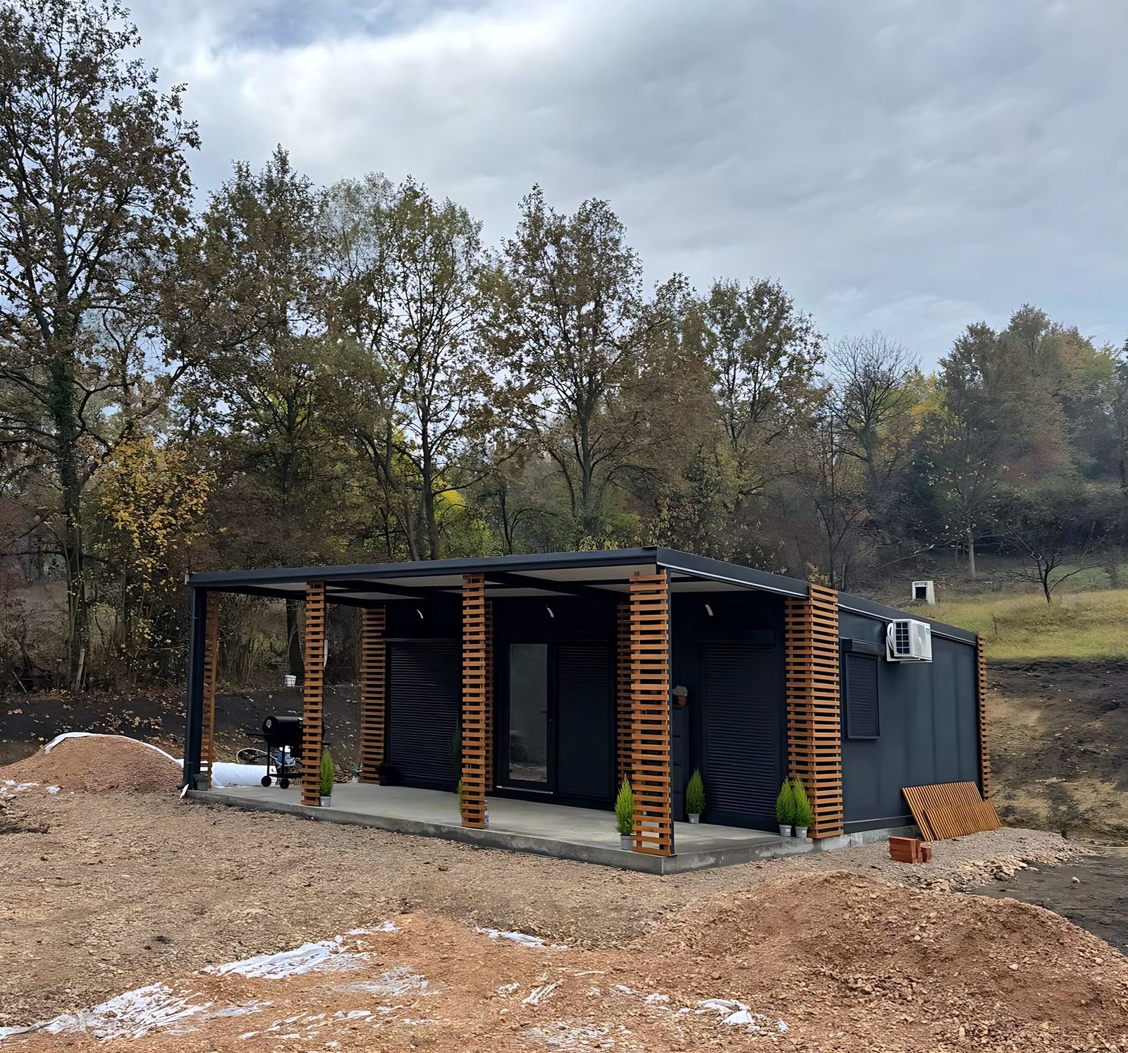 Antracit színű modulház fedett terasszal és falamellákkal, őszi erdő előtt – az Ecohouse, a GVM Haus gyártópartnere mintaháza